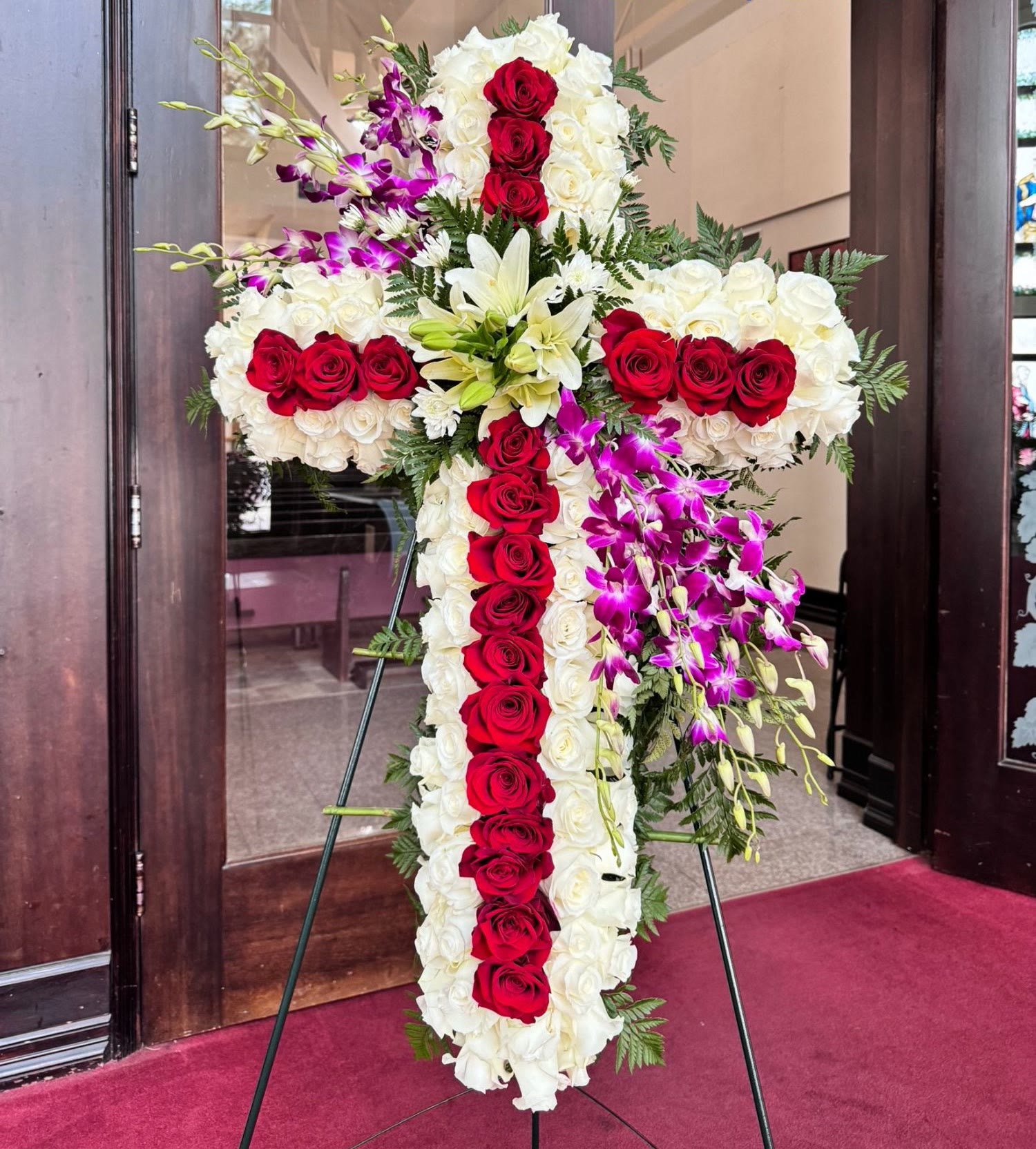 A standing flower cross of white roses with a line of red roses, white lilies, and purple orchids, in front of a church door.