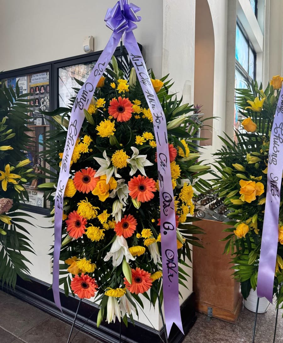 Standing spray of orange gerberas, yellow mums, and white lilies with a lavender ribbon.